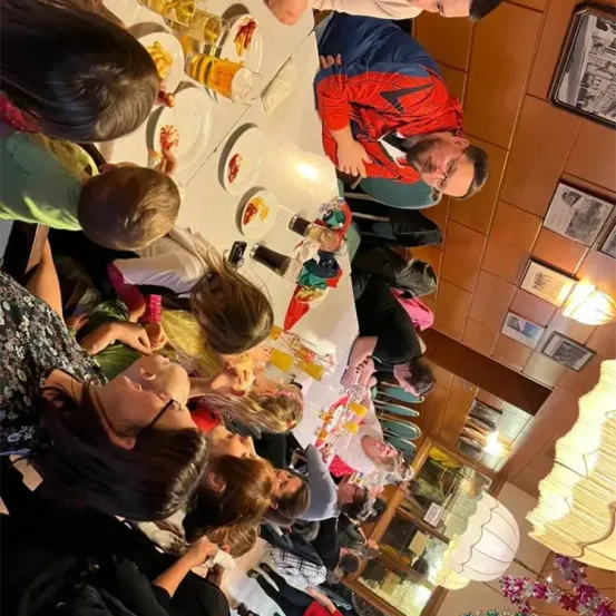 Mehrere Personen sitzen an einem langen Tisch mit Essen, Getränken und Dekorationen. Eine Person in einer Spider-Man-Kostüm beugt sich vor.