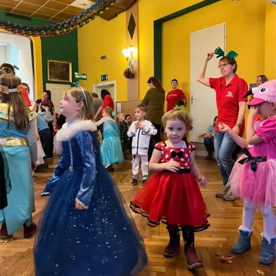 Eine Gruppe von Kindern in Kostümen tanzt in einem Raum. Ein Kind trägt ein Minnie-Maus-Kostüm. Ein Erwachsener steht mit einer grünen Papageienmütze hinter ihnen. Der Raum hat einen Holzboden, gelbe und grüne Wände und Dekorationen.