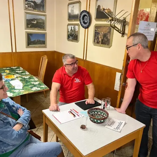 Drei Personen in einem Restaurant, zwei Männer in roten Shirts, eine Frau in blauer Jeans. Ein Tisch hat eine Schüssel, Stifte und ein Papier mit 'Freie Spende'. Dahinter Stühle und ein Tisch mit blumiger Tischdecke. Die Wand hat mehrere gerahmte Bilder.