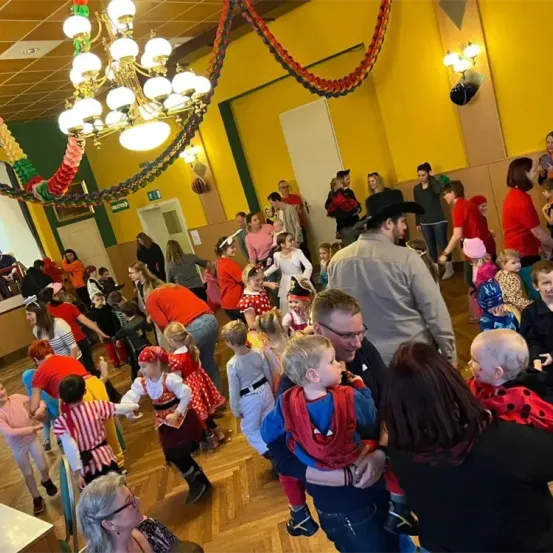 Eine Gruppe von Menschen in Kostümen und mit Brillen ist in einem Saal, wahrscheinlich für ein Festival. Der Saal ist mit einer Kronleuchter, bunten Bändern und Lichtern dekoriert. Einige Menschen halten Kinder.