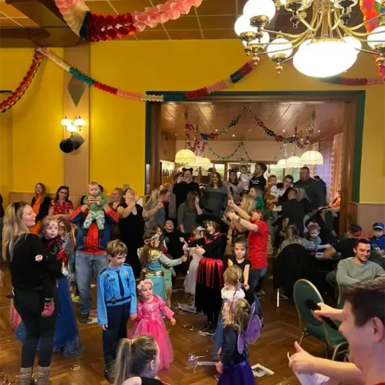 Eine festliche Zusammenkunft mit vielen Menschen in einem Partysaal. Mehrere Personen sind in Kostümen gekleidet. Eine Kronleuchter und Lampen hängen von der Decke. Bunte Dekorationen schmücken die Wände.