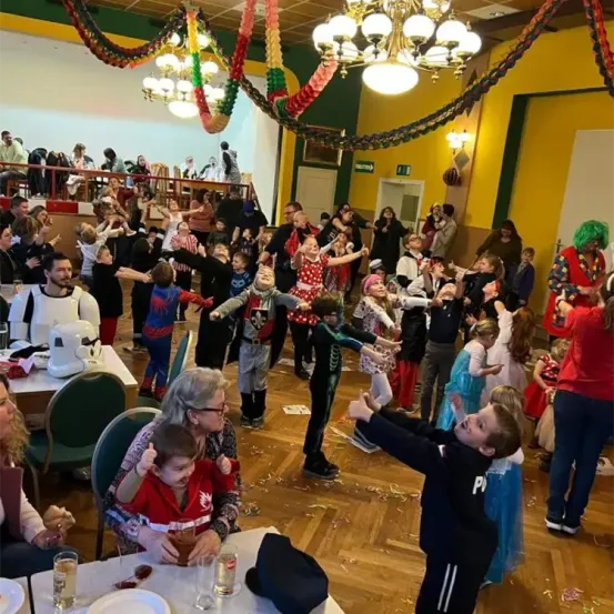 Eine festliche Zusammenkunft mit vielen Kindern in Kostümen, die tanzen und spielen. Eine Frau hält ein Kind in einem roten Kostüm. Menschen stehen und sitzen um Tische mit Getränken und Essen. Eine Kronleuchter und bunte Dekorationen schmücken den Raum.