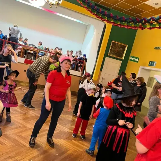 Eine Gruppe von Kindern und Erwachsenen ist drinnen, einige sind in Kostümen gekleidet. Eine Frau in Rot leitet einen Tanz. Ein Schild heißt 'Toilette' und ein anderes weist auf eine Toilette hin.