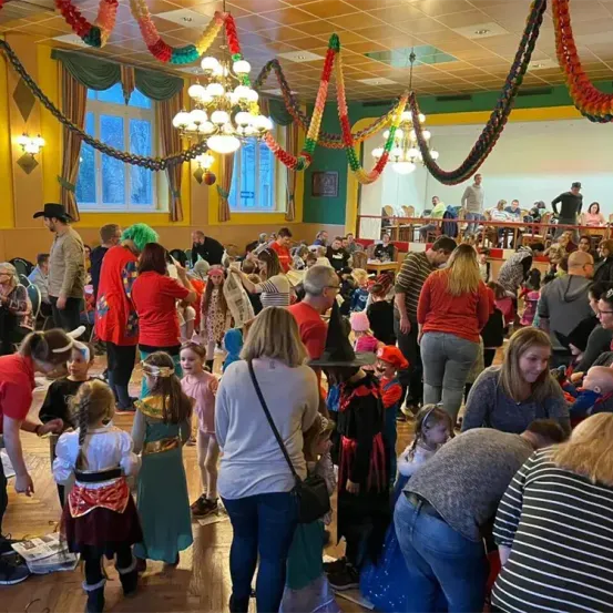 Ein Saal voller Menschen, einige in Kostümen, andere in Freizeitkleidung. Ein Kind verkleidet als Pirat und ein anderes als Hexe. Zwei Kronleuchter hängen von der Decke.