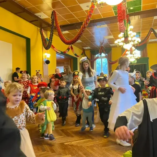 Eine Gruppe von Kindern in Kostümen tanzt in einem dekorierten Raum. Zwei Frauen führen den Tanz an, eine in einem weißen Kleid und die andere in einem gestreiften Kleid.
