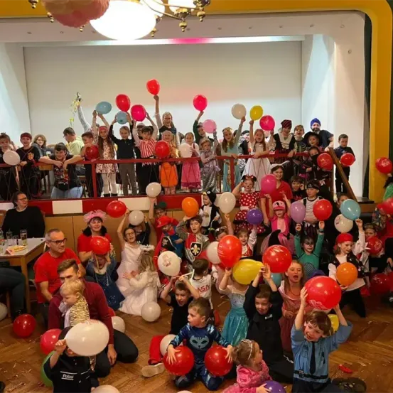 Eine große Gruppe von Kindern und Erwachsenen versammelt sich in einem Raum, viele halten bunte Luftballons. Einige stehen auf dem Boden, andere sitzen. Der Raum ist mit Luftballons und Girlanden dekoriert, und es herrscht eine festliche Atmosphäre.