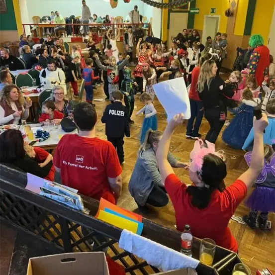 Ein Saal voller Menschen, hauptsächlich Kinder, in Kostümen und festlichen Dekorationen. Erwachsene und Kinder beschäftigen sich mit Aktivitäten, einige stehen und halten Papiere. Der Boden ist mit bunten Gegenständen dekoriert.