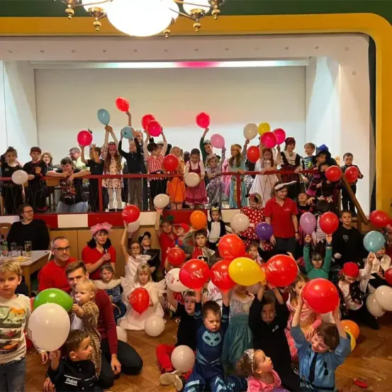 Eine große Gruppe von Kindern und Erwachsenen versammelt sich in einem Raum und hält bunte Luftballons hoch. Sie sind in verschiedenen Kostümen gekleidet, einige mit Gesichtsbemalung. Ein paar Erwachsene stehen hinter einem Geländer, und eine Kronleuchter hängt von der Decke.