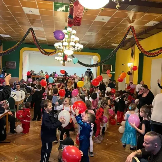 In einem festlichen Saal feiern Kinder und Erwachsene. Der Raum ist gefüllt mit bunten Luftballons und Konfetti. Die Kinder halten Ballons, während einige Erwachsene Kameras halten und den Moment festhalten. Der Saal hat einen Holzboden, grün und gelbe Wände und eine Kronleuchter, die von der Decke hängt.