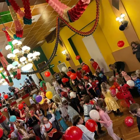Eine große Gruppe von Kindern und Erwachsenen feiert ein festliches Ereignis in Innenräumen. Der Raum ist mit Ballons, Luftschlangen und bunten Lichtern geschmückt. Die Kinder halten Ballons und tragen Kostüme, während einige Erwachsene ihnen helfen.