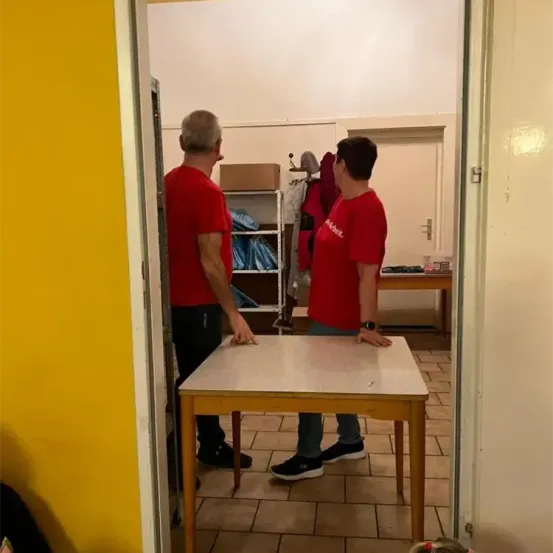 Zwei Männer in roten Shirts stehen an einem Tisch, einer scheint zu gestikulieren. Dahinter führt eine Tür in einen Raum mit Regalen und einem Tisch.