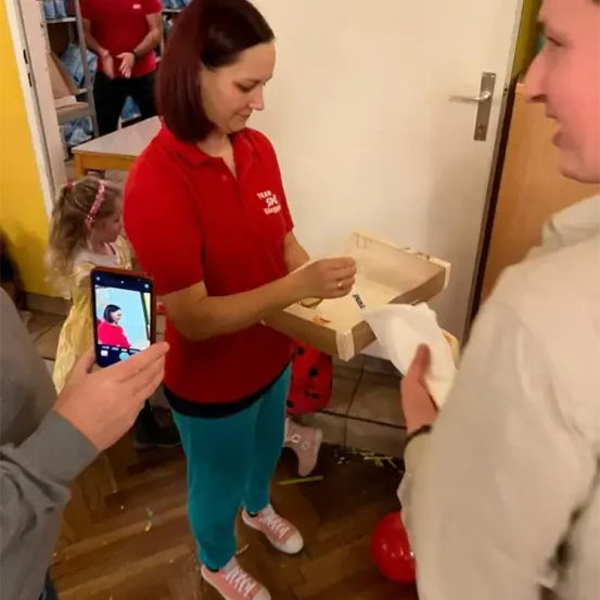 Eine Frau in Rot hält eine Pizzaschachtel und gibt sie einem Mann, während ein Kind und eine weitere Person zusehen. Ein Smartphone hält den Moment fest.