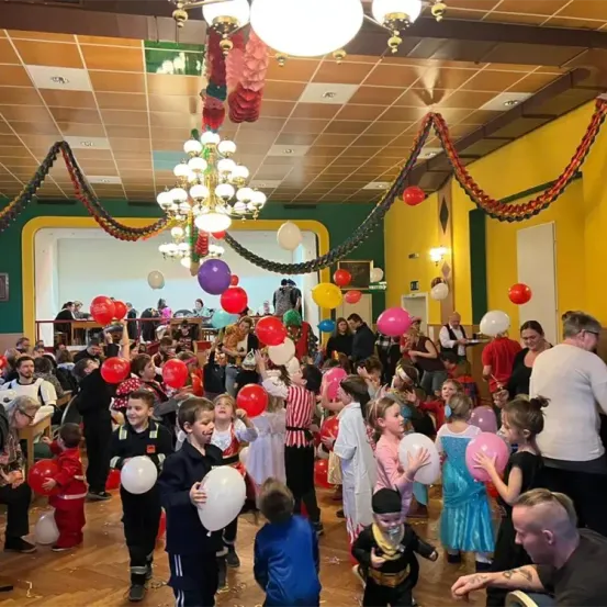Eine große Gruppe von Kindern in verschiedenen Kostümen befindet sich in einem Raum voller Luftballons. Der Raum hat eine festliche Atmosphäre mit bunten Dekorationen, die von der Decke und den Wänden hängen. Einige Erwachsene sind ebenfalls anwesend, entweder als Zuschauer oder Teilnehmer.