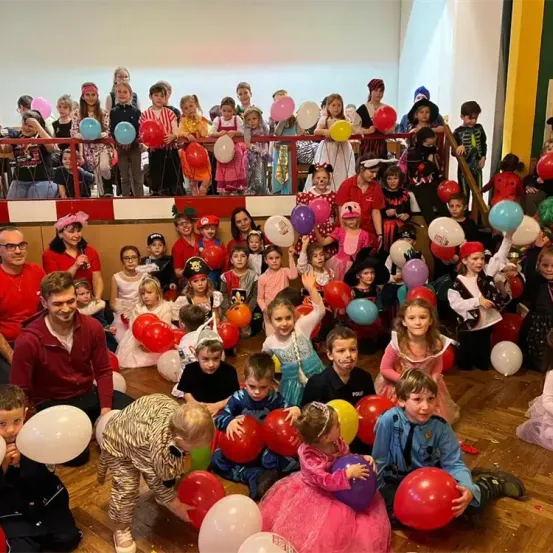 Eine Gruppe von Kindern in verschiedenen Kostümen mit Luftballons, sitzend und stehend auf einem Holzboden, mit Erwachsenen und weiteren Kindern im Hintergrund.