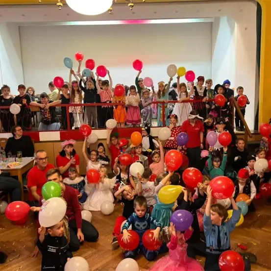 Eine Gruppe von Kindern in Kostümen und Erwachsenen feiert in einem Saal mit Luftballons und einer Bühne. Einige sind auf dem Boden, während andere auf einer Balkon sind. Der Saal hat Holzböden und rote Geländer.