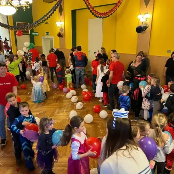 Eine Gruppe von Kindern in Kostümen und Erwachsenen versammelt sich in einem dekorierten Raum voller Luftballons. Einige Kinder halten Luftballons, während andere stehen und zusehen. Der Raum hat gelbe Wände und eine Kronleuchter.