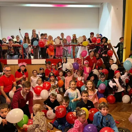 Eine Gruppe von Kindern und Erwachsenen versammelt sich in einem Raum und hält bunte Luftballons. Einige Kinder sind in Kostümen gekleidet, und Erwachsene lächeln und posieren für ein Foto. Der Raum hat eine weiße Wand, ein rotes Geländer und einen Holzboden.