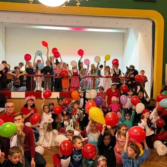 Eine große Gruppe von Kindern und Erwachsenen in Kostümen versammelt sich, bunte Luftballons haltend, in einem festlichen Raum mit Bühne und gelber Brüstung.