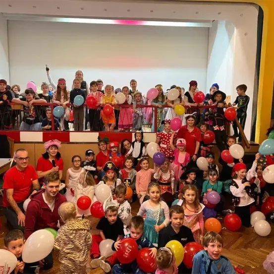 Eine große Gruppe von Kindern und Erwachsenen in Kostümen, die bunte Luftballons halten und für ein Foto posieren. Erwachsene stehen auf dem Balkon und beobachten die Szene.