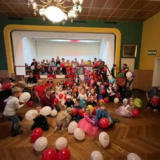 Eine große Gruppe von Kindern in Kostümen versammelt sich in einem Saal mit Luftballons. Sie sitzen und stehen auf dem Boden, während Erwachsene in der Nähe stehen. Eine Kronleuchter hängt von der Decke.
