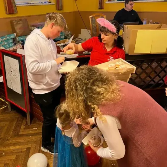 Ein Mann in einer weißen Kapuzenjacke reicht einer Mädchen in einer rosa Mütze eine Backware. Eine weitere Frau und ein Mädchen betrachten eine Schachtel mit Gebäck. Im Hintergrund sitzt eine Person an einem Tisch.