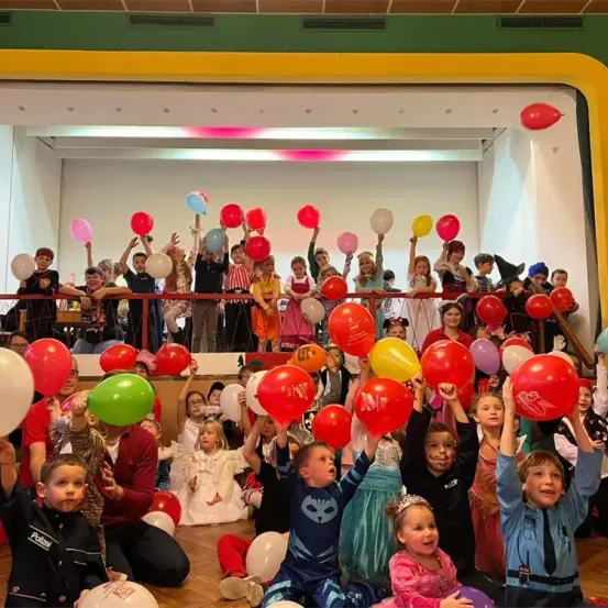 Eine Gruppe von Kindern und Erwachsenen in festlicher Kleidung versammelt sich in einem Raum und hält Luftballons in verschiedenen Farben und Größen hoch. Einige sitzen, andere stehen auf den Stufen. Eine festliche Atmosphäre erfüllt den Raum.