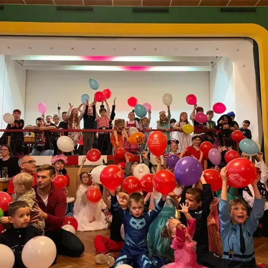 Kinder und Erwachsene in einem Saal, halten und werfen Luftballons, einige in Kostümen, in einer festlichen Atmosphäre mit bunten Luftballons und Leuten auf einem Balkon darüber.