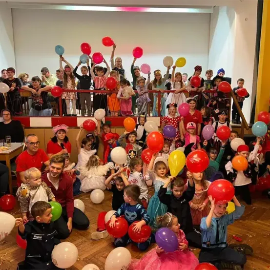 Eine Gruppe von Menschen, einschließlich Kindern und Erwachsenen, versammelt sich in einem Saal, viele halten bunte Luftballons. Einige sitzen, während andere auf einer Balkon stehen. Die Umgebung wirkt festlich.