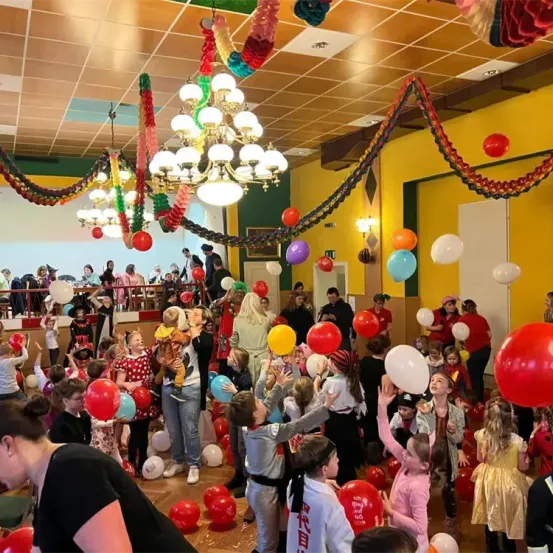 Ein großer Raum mit gelber Wand ist gefüllt mit vielen Menschen, hauptsächlich Kindern, die mit Luftballons in verschiedenen Farben spielen. Dekorationen hängen von der Decke, und eine Frau hält ein Kind hoch.