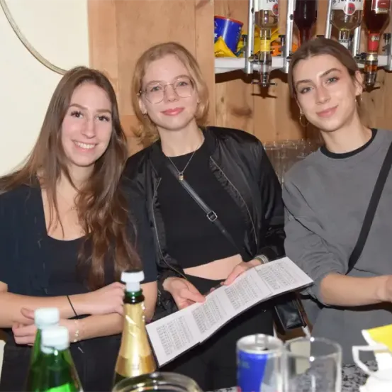 Drei Frauen stehen hinter einer Bartheke, lächeln und posieren für ein Foto. Die Frau links hat lange Haare und trägt ein schwarzes Oberteil. Die Frau in der Mitte trägt eine Brille und eine schwarze Jacke. Die Frau rechts trägt ein graues Oberteil und eine schwarze Schultertasche.