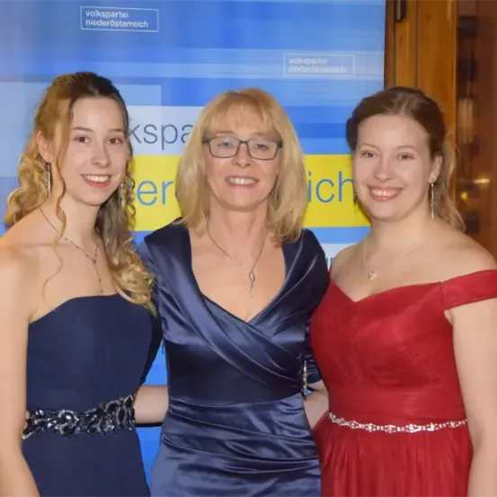 Drei Frauen stehen nebeneinander, wahrscheinlich bei einer Veranstaltung, in Abendkleidern. Die Frau in der Mitte trägt eine Brille. Hinter ihnen ist ein Hintergrund mit Text und Logos.