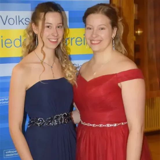 Zwei Frauen lächeln und posieren für ein Foto vor einem Schild mit blauem und gelbem Hintergrund. Die Frau auf der linken Seite trägt ein blaues Kleid und eine Halskette. Die Frau auf der rechten Seite trägt ein rotes Kleid und eine Halskette.