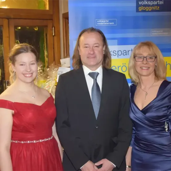 Drei Menschen stehen eng beieinander und lächeln bei einer Veranstaltung. Eine Frau in Rot, ein Mann in einem Anzug und eine weitere Frau in Blau. Hinter ihnen steht ein Banner mit der Aufschrift 'volkspartei gloggnitz'.