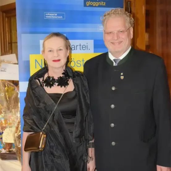 Ein Mann und eine Frau, beide in Schwarz gekleidet, lächeln und stehen eng nebeneinander vor einem Plakat.