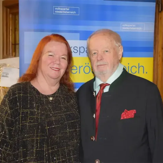 Eine Frau mit roten Haaren und einem schwarzen karierten Oberteil steht neben einem Mann in einem schwarzen Anzug mit roter Krawatte und Einstecktuch. Sie stehen vor einem blau-gelben Banner mit deutschem Text.
