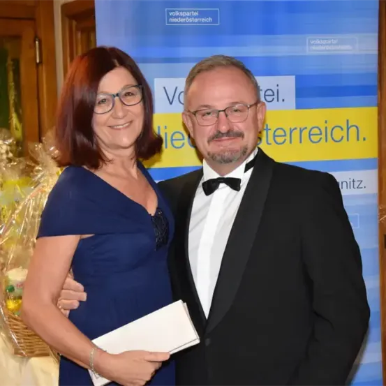 Ein Mann und eine Frau in formeller Kleidung posieren für ein Foto vor einem blauen Hintergrund mit Text. Die Frau hält eine Mappe und trägt eine Brille. Dahinter steht ein Blumenkorb.