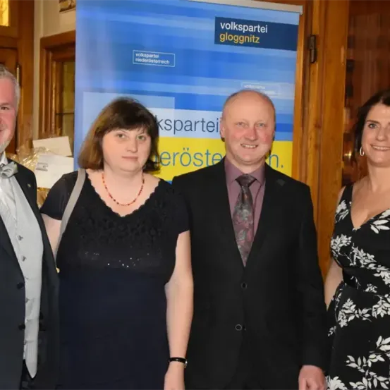 Vier Erwachsene stehen vor einem blauen Banner mit gelbem Text. Der Mann auf der linken Seite trägt eine Fliege. Die Frau in der Mitte trägt eine Halskette. Der Mann neben ihr trägt eine Krawatte. Die Frau auf der rechten Seite trägt ein schwarz-weißes Kleid.