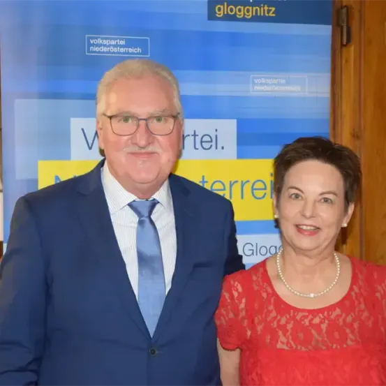 Ein älterer Mann und eine ältere Frau stehen nebeneinander vor einem blau-gelben Schild mit Text. Der Mann trägt einen Anzug und eine Krawatte, die Frau ein rotes Kleid und eine Perlenkette.