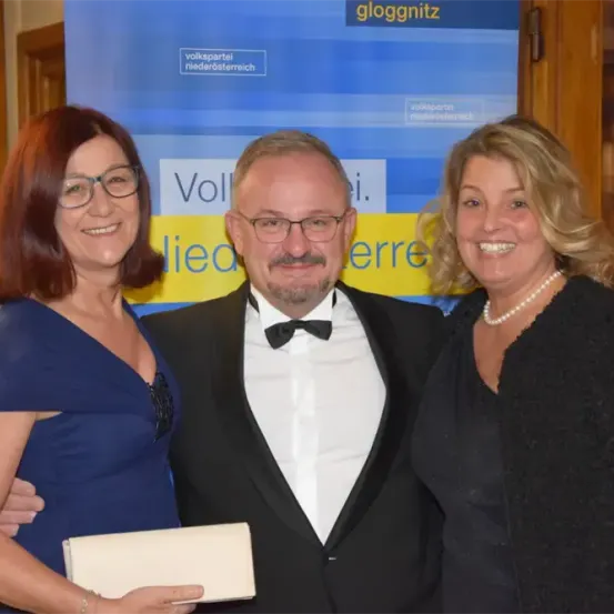 Drei Menschen posieren für ein Foto. Zwei Frauen und ein Mann. Sie sind in Abendgarderobe gekleidet. Der Mann trägt einen Fliege. Sie lächeln für die Kamera. Der Hintergrund ist ein Banner mit Text.