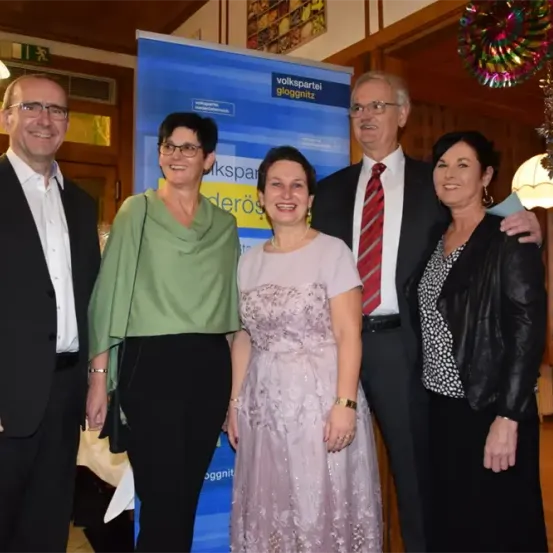 Fünf Menschen stehen gemeinsam für ein Foto in einem Raum. Sie alle lächeln und posieren für die Kamera. Hinter ihnen ist ein Banner mit Text und einer Lampe.