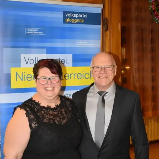 Ein lächelndes Paar steht vor einem blauen Banner mit Text auf Deutsch. Der Mann trägt einen Anzug und eine Krawatte, und die Frau trägt ein schwarzes Kleid und eine Brille. Sie lächeln beide.