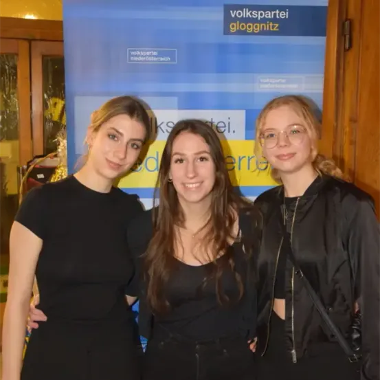 Drei Frauen stehen eng beieinander vor einem blauen Banner mit dem Text 'volkspartei gloggnitz'. Sie tragen alle schwarze Kleidung und scheinen für ein Foto zu lächeln.