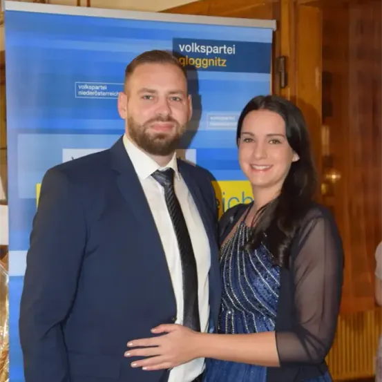 Ein Mann und eine Frau stehen eng beieinander vor einem blauen Banner, auf dem 'volkspartei gloggnitz' steht. Der Mann trägt einen Anzug und eine Krawatte, die Frau ein blaues Kleid. Sie lächeln beide in die Kamera.