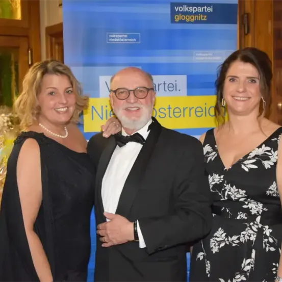 Drei Personen stehen lächelnd für ein Foto zusammen. Ein Mann im Smoking steht zwischen zwei Frauen, die beide schwarze Kleider tragen. Hinter ihnen befindet sich ein blauer Banner mit weißem Text, der 'Volkspartei Niederosterreich' lautet.