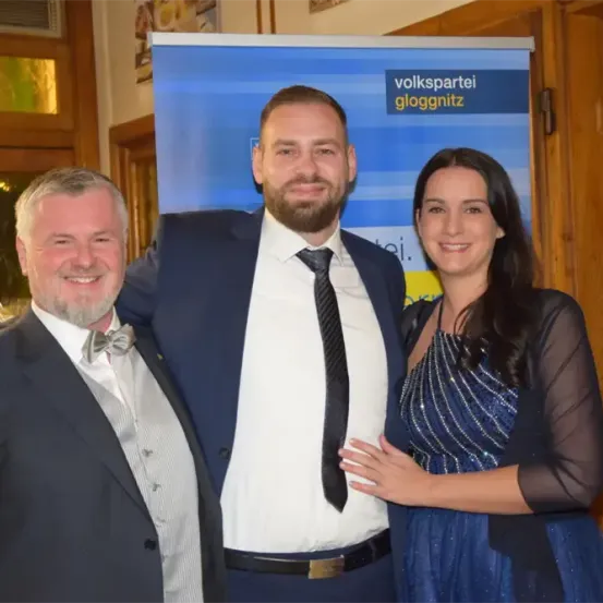 Drei Menschen posieren für ein Foto bei einer Veranstaltung. Ein Mann in formeller Kleidung und eine Frau in einem blauen Kleid stehen neben einem Mann in Anzug und Krawatte. Hinter ihnen befindet sich ein blauer Banner mit dem Text 'volkspartei gloggnitz'.