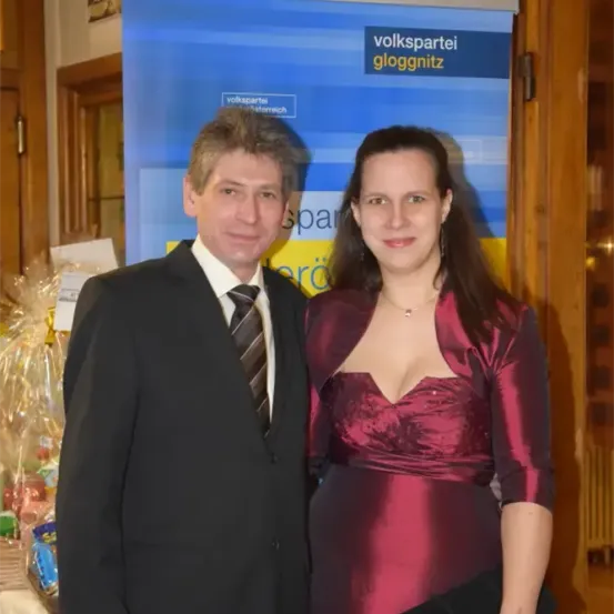 Ein Mann und eine Frau stehen nebeneinander vor einem Banner mit der Aufschrift 'volkspartei gloggnitz'. Der Mann trägt einen schwarzen Anzug und eine Krawatte, und die Frau trägt ein weinrotes Kleid. Dahinter befindet sich eine Holztür und eine weiße Tischdecke mit Gegenständen darauf.