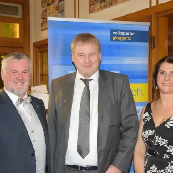 Drei Personen stehen gemeinsam für ein Foto; ein Mann links trägt eine Schleife, ein Mann in der Mitte trägt einen grauen Anzug und eine Krawatte und eine Frau rechts lächelt. Hinter ihnen befindet sich ein blauer Banner mit 'volkspartei gloggnitz' darauf.