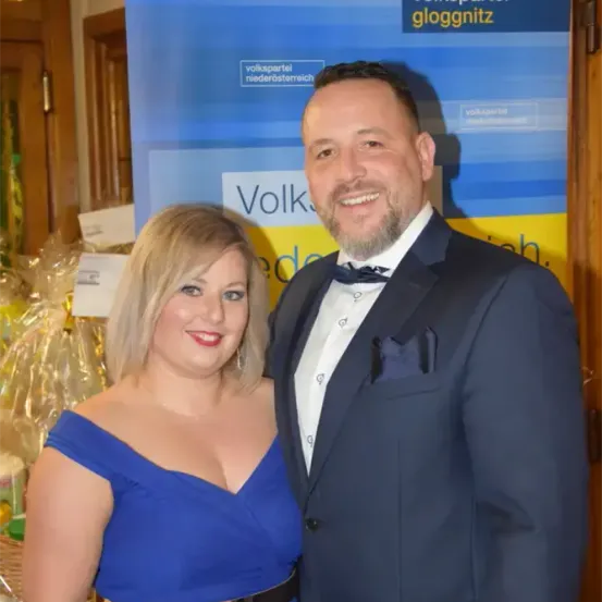 Ein Mann und eine Frau in formeller Kleidung lächeln für die Kamera vor einem Volkspartei Niederosterreic-Schild.