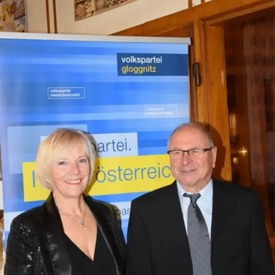 Ein Mann und eine Frau stehen neben einem Volkspartei Gloggnitz-Banner. Die Frau trägt eine Halskette und der Mann trägt eine Brille und eine Krawatte.