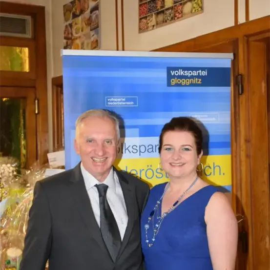 Ein älterer Mann und eine Frau lächeln und stehen neben einem Banner für die Volkspartei Gloggnitz.